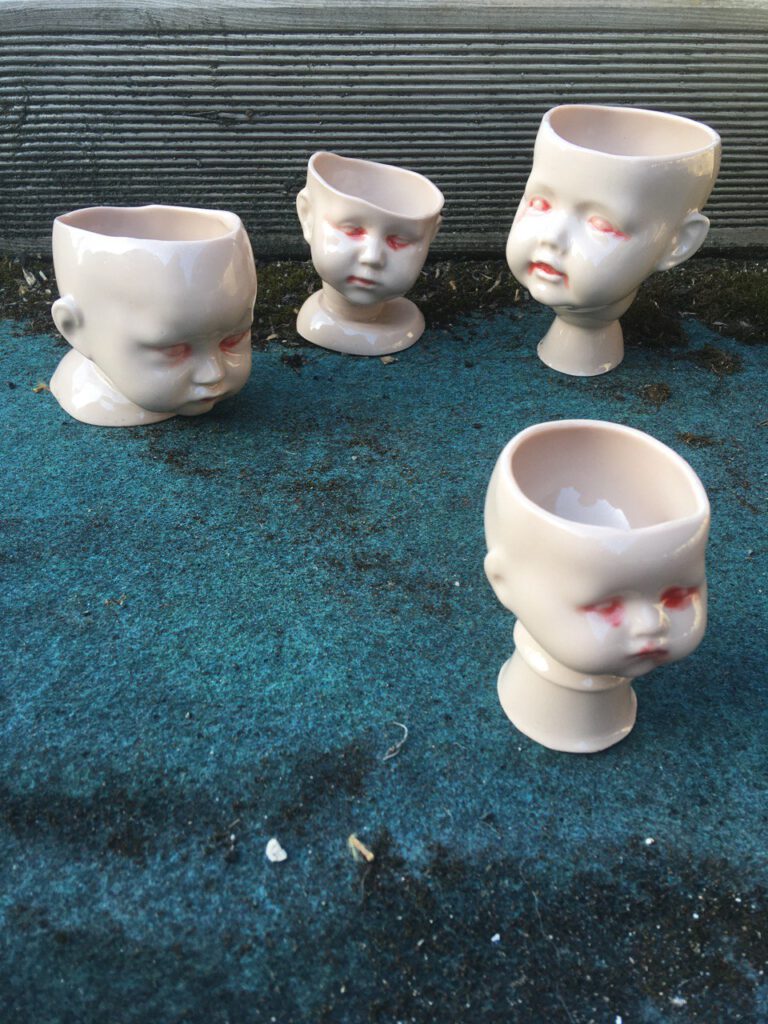 Four white ceramic cups, that look like baby heads are staning in a green carpet. The eyes of the baby-cups look bloodshot.